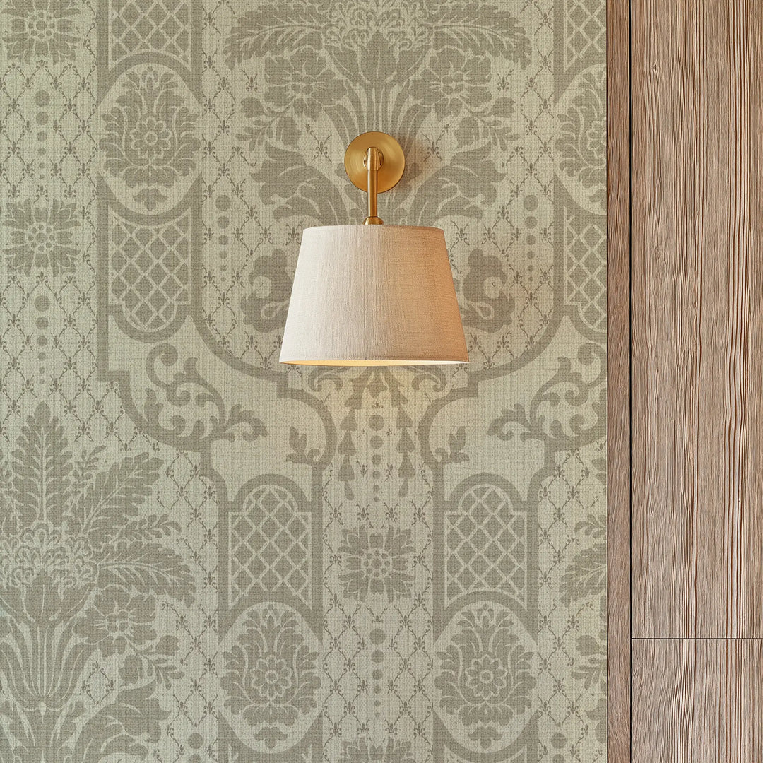 A wall-mounted sconce with a beige fabric drum shade and a gold-finished metal mount, shown against a patterned wallpaper background.