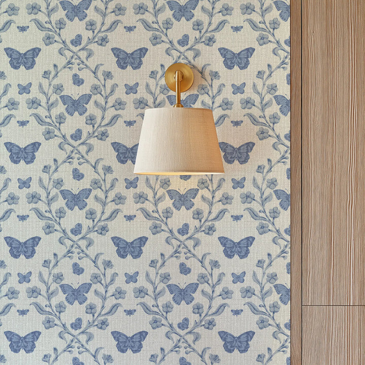 A wall-mounted sconce with a beige fabric drum shade and a gold-finished wall mount, shown against wallpaper with a repeating blue butterfly and floral pattern and a light wood panel on the right.