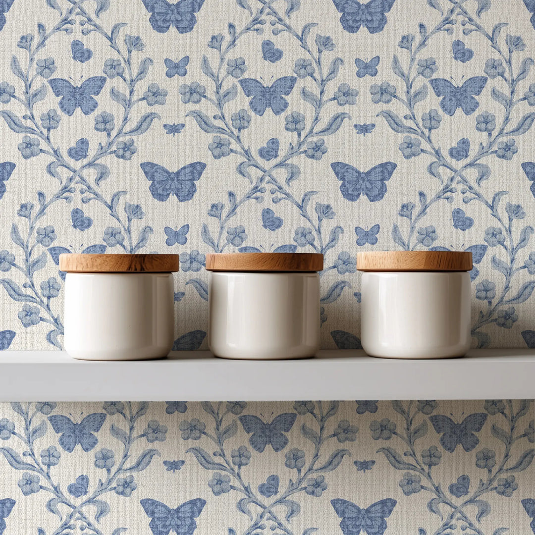 Three cream-colored ceramic canisters with light wood lids are arranged on a white shelf against a blue and white butterfly patterned wallpaper background.